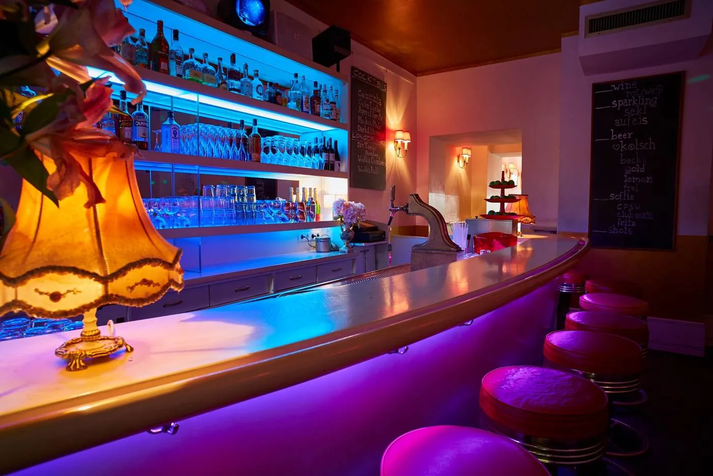 Hotelbar im Maison Marsil mit geschwungenem Tresen, beleuchteten Regalen und roten Barhockern