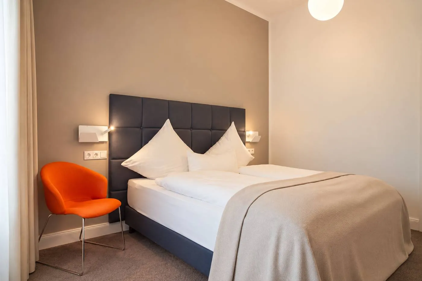 Zimmer mit Doppelbett, grauem Kopfteil und orangem Sessel